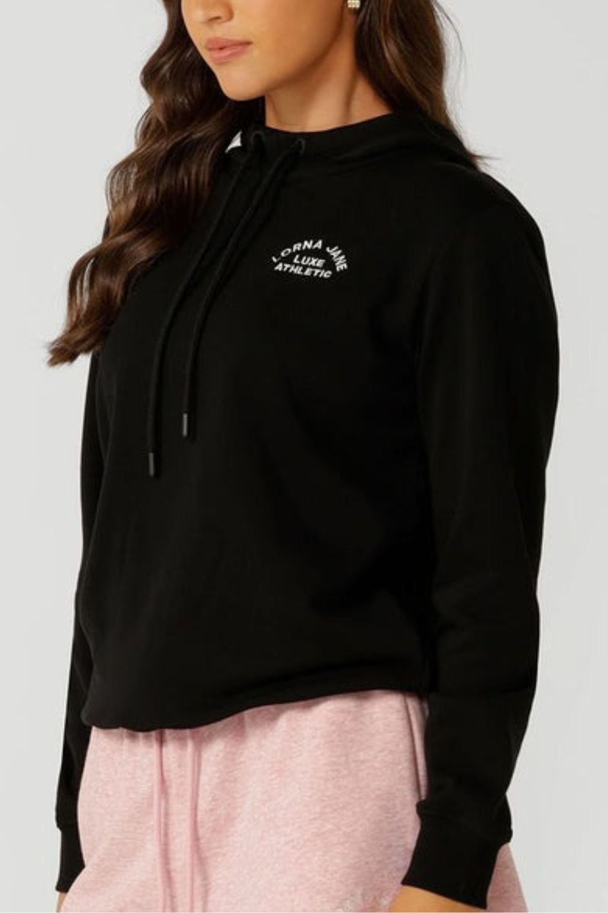 A woman is wearing the Lorna Jane Lotus Hoodie in black with white chest text and light pink sweatpants, standing against a light gray background. Only her upper body is visible. The hoodie is made from a breathable cotton blend.
