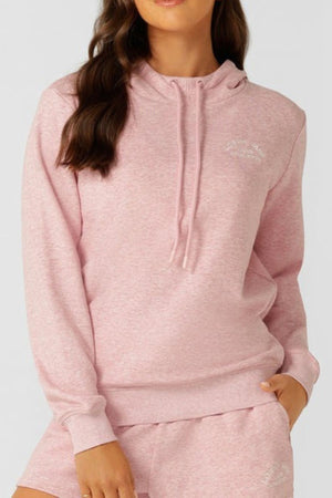 A woman wears the Lorna Jane Lotus Hoodie in light pink—a relaxed fit, cotton-blend terry sweatshirt with a drawstring hood, subtle chest text, and matching shorts—standing with one hand in her pocket.