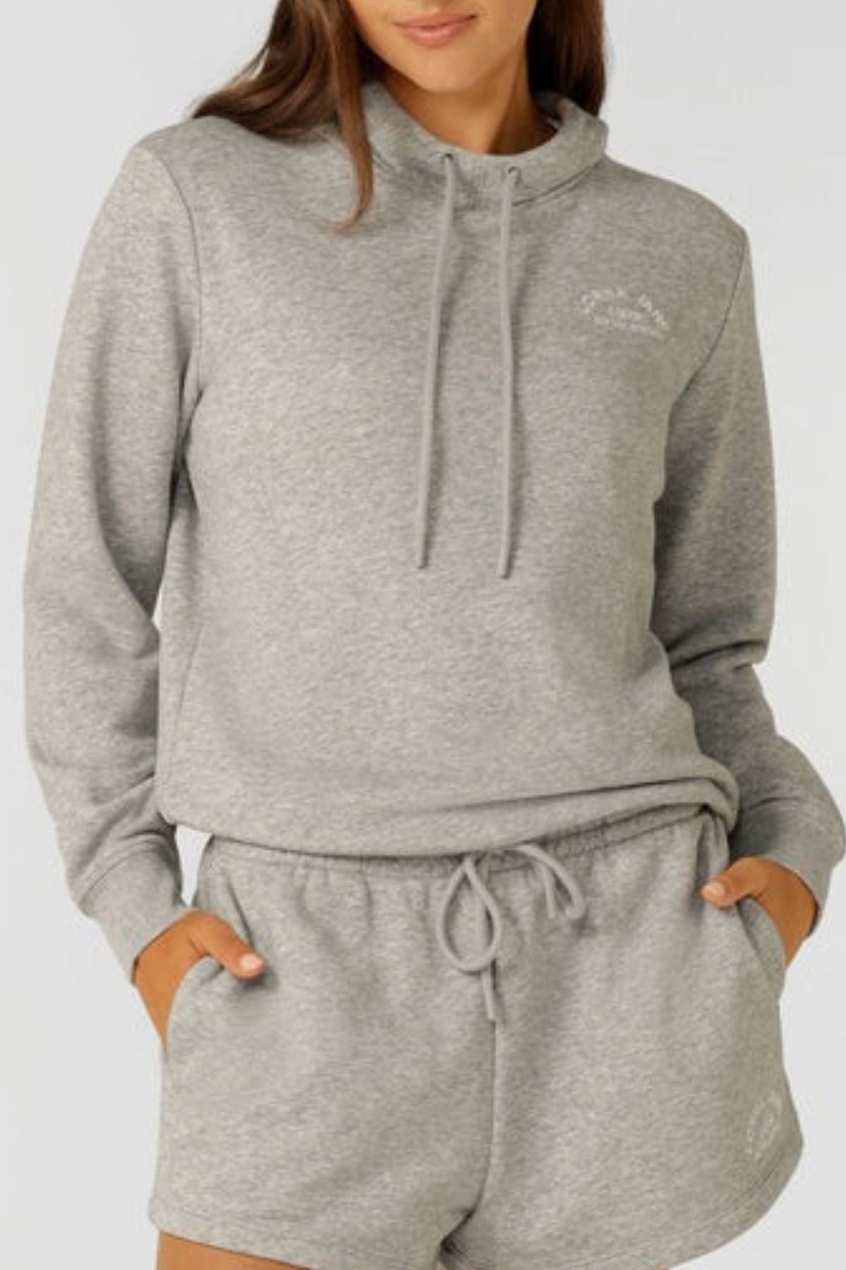 A woman wearing the Lorna Jane Lotus Hoodie in light gray cotton blend with drawstrings and matching gray shorts, standing with her hands in her pockets against a plain background.