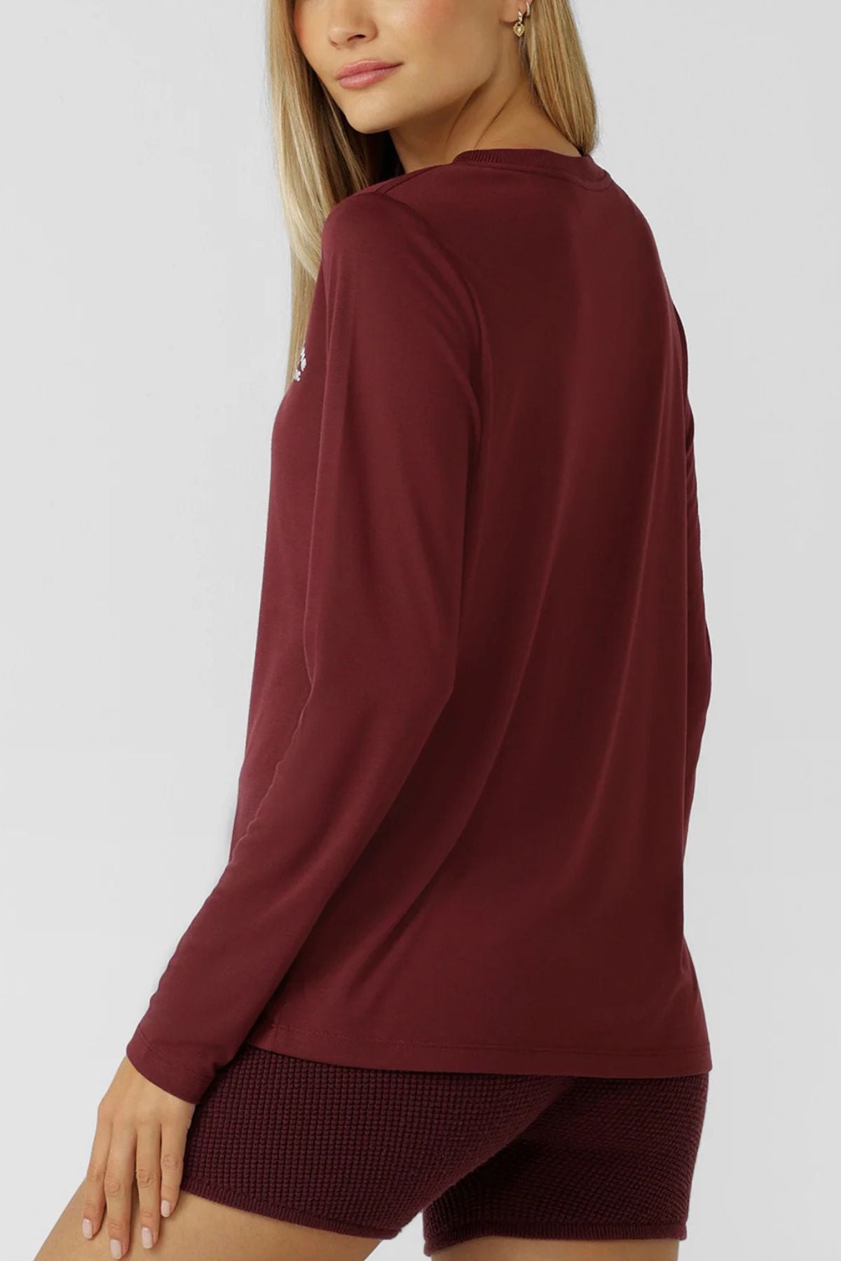A woman with long blonde hair wears the Lorna Jane Lotus Long Sleeve T-Shirt and textured maroon shorts, posing with her back slightly turned and looking over her shoulder. The activewear is made from lightweight, breathable fabric against a plain light background.