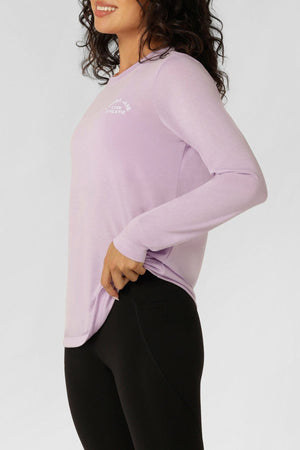 Woman in a Lorna Jane Lotus Long Sleeve T-Shirt and black leggings stands sideways, smiling against a plain light background—showcasing stylish activewear with a lightweight, breathable long sleeve top.