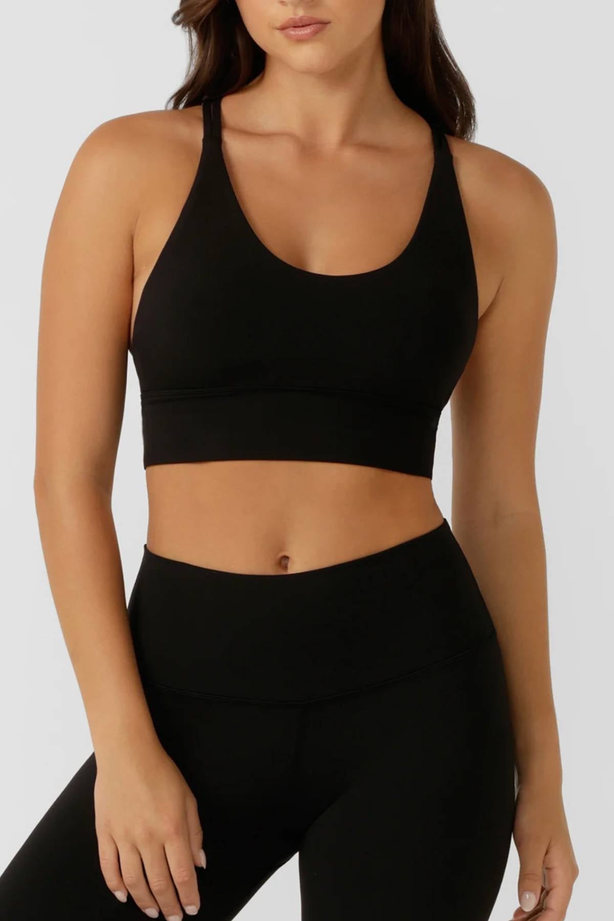 A woman wears the Lorna Jane Lotus Longline Sports Bra and high-waisted black leggings, standing against a plain light gray background with her head partially out of frame.