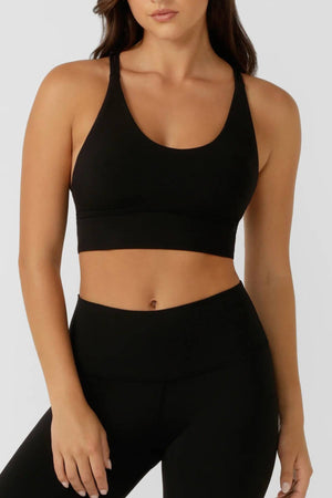 A woman wears the Lorna Jane Lotus Longline Sports Bra and high-waisted black leggings, standing against a plain light gray background with her head partially out of frame.