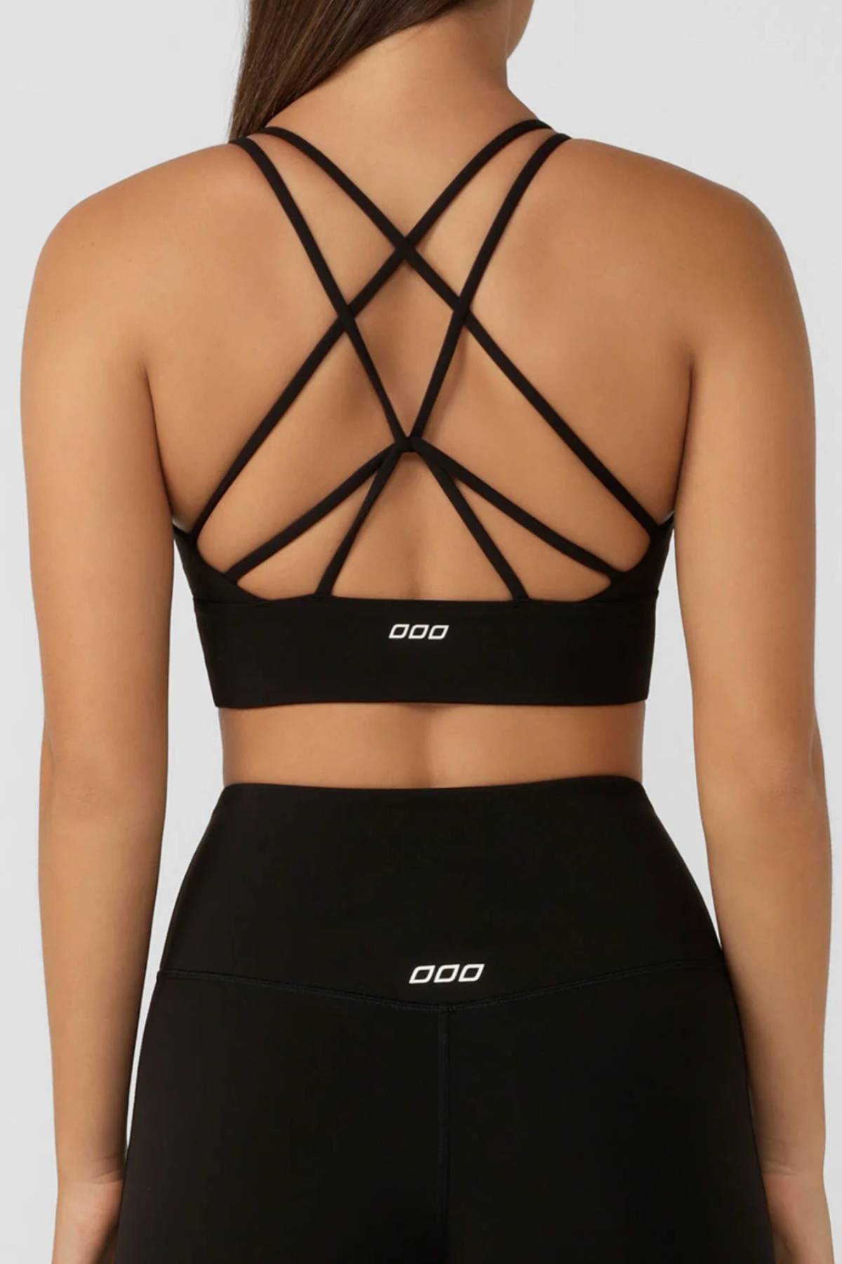 A woman wears the Lorna Jane Lotus Longline Sports Bra in black, featuring moisture-wicking fabric and multiple crisscross back straps, paired with matching high-waisted leggings. Both have a small white logo and are shown from the back for style and comfort.