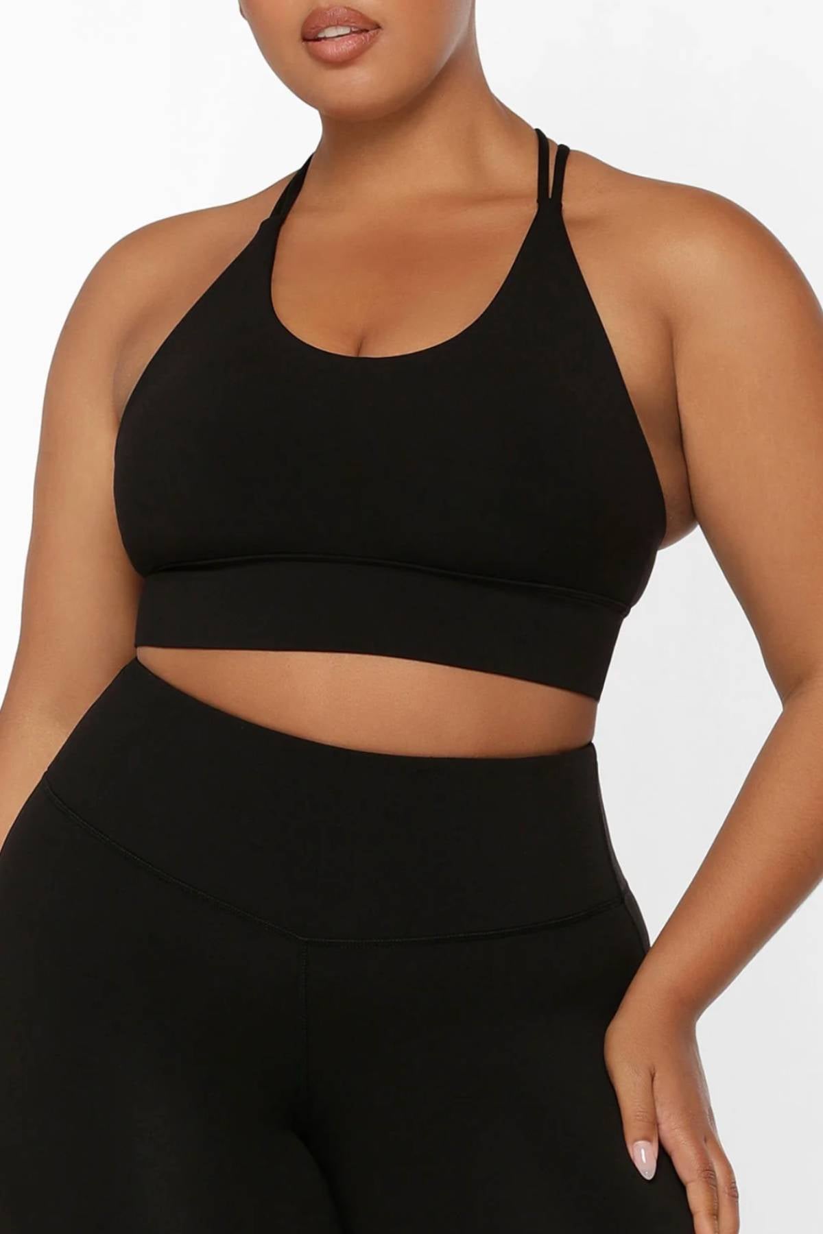A woman in the Lorna Jane Lotus Longline Sports Bra and high-waisted black leggings stands against a white background, her right hand on her hip. Her face is partially visible. The outfit offers 4-way stretch for comfort and fit.