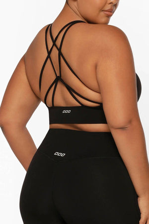 A woman is shown from behind on a white background, wearing the Lorna Jane Lotus Longline Sports Bra in black with multiple crisscross back straps and matching black leggings.
