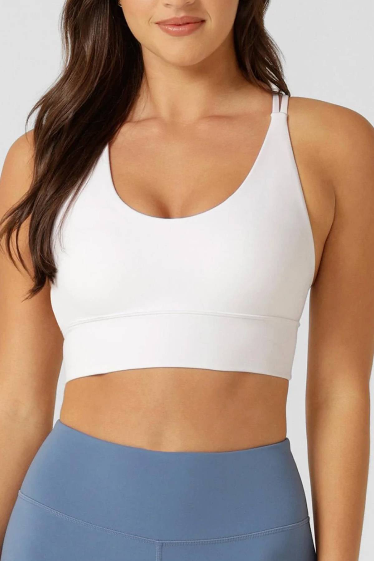 A woman in a Lorna Jane Lotus Longline Sports Bra and high-waisted blue leggings stands against a plain light gray background.