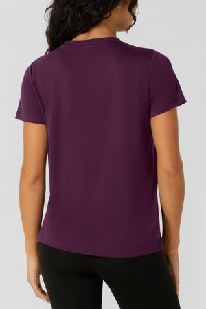 A woman with wavy dark hair wears the Lorna Jane Lotus T-Shirt in purple and black pants, standing with her back to the camera against a plain light background.