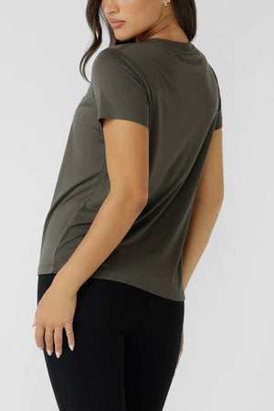 A woman stands slightly turned away, wearing the Lorna Jane Lotus T-Shirt in olive green with black leggings against a plain light background. Her hair is loose, and her left arm rests by her side.