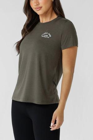 A woman wears the Lorna Jane Lotus T-Shirt, a women's olive green crew neck tee with "Lorna Jane Active Athletic" on the chest, paired with black leggings, standing against a light background.