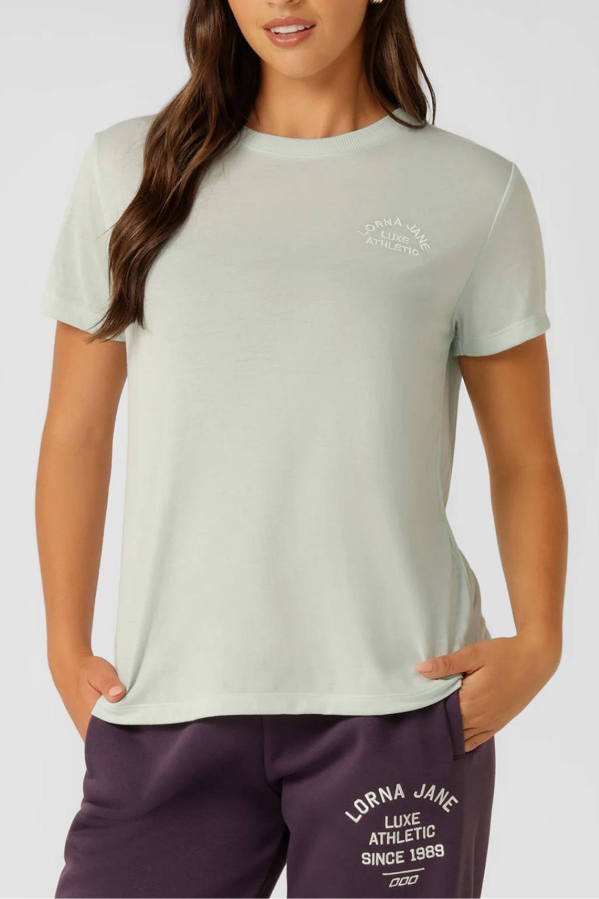 A woman embraces an active lifestyle in a light sage green Lorna Jane Lotus T-Shirt with "Lorna Jane Luxe Athletic" on the chest, paired with dark purple sweatpants with a white logo near the pocket.