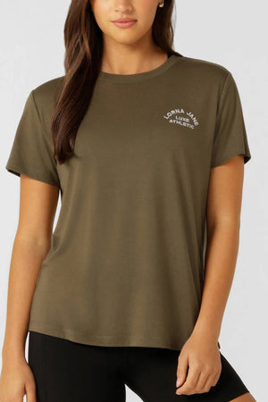 A woman with long brown hair wears the olive-green Lorna Jane Lotus T-Shirt, featuring "Lorna Jane Luxe Athletic" in white on the left chest, creating a classic active look against a plain background.