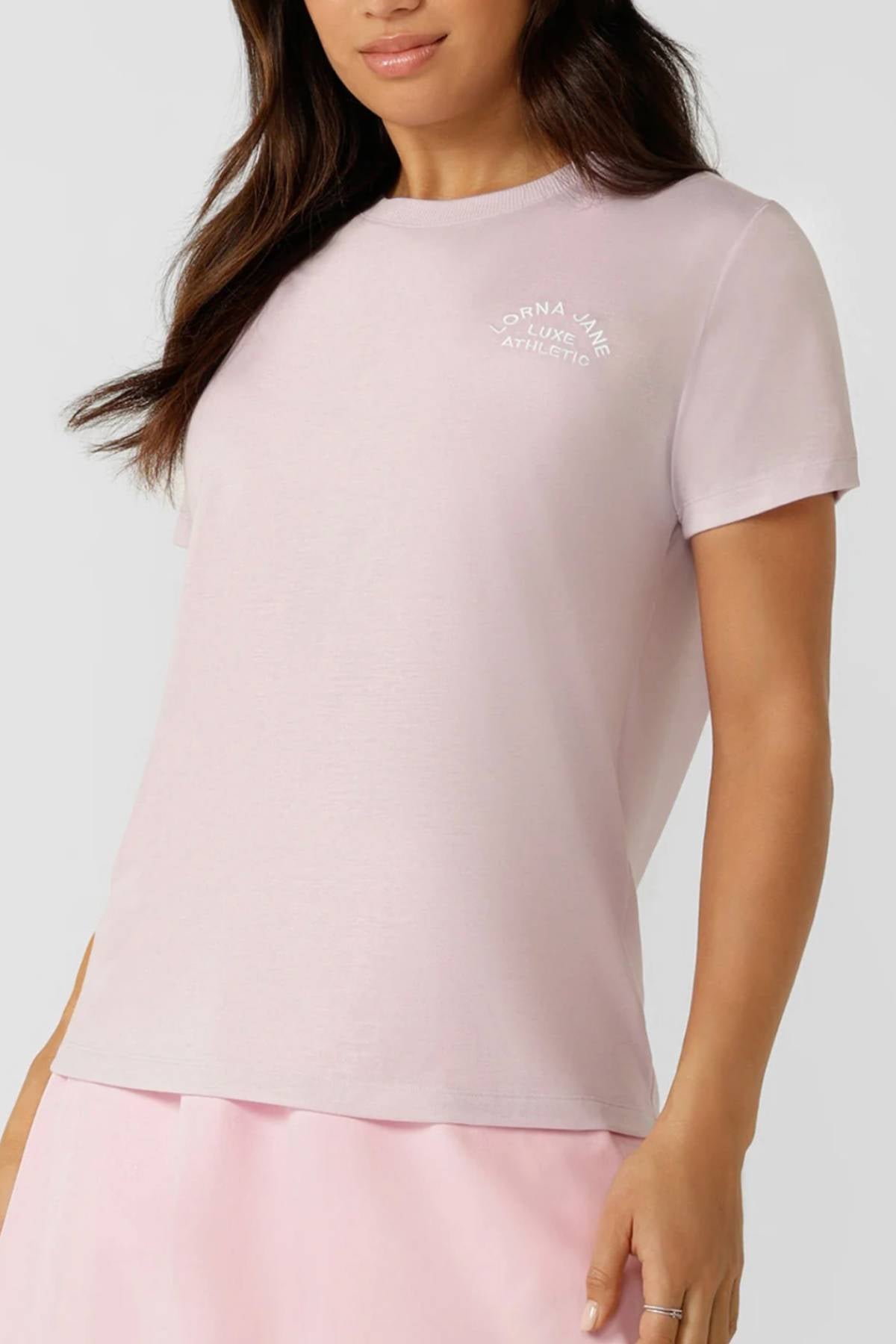 Wearing a light pink Lorna Jane Lotus T-Shirt with an embroidered logo and "True Athletic" text on the chest, paired with a matching skirt, she showcases an active lifestyle against a simple background.