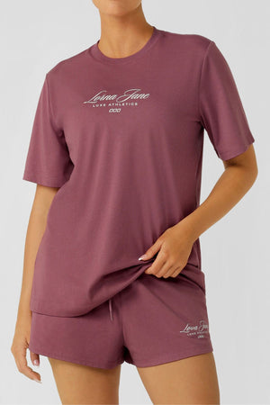 A person wears a matching mauve Lorna Jane Luxe Athletic Relaxed T-Shirt and shorts set in breathable cotton blend with "Lorna Jane Luxe Athletics" printed in white, standing against a plain light background.