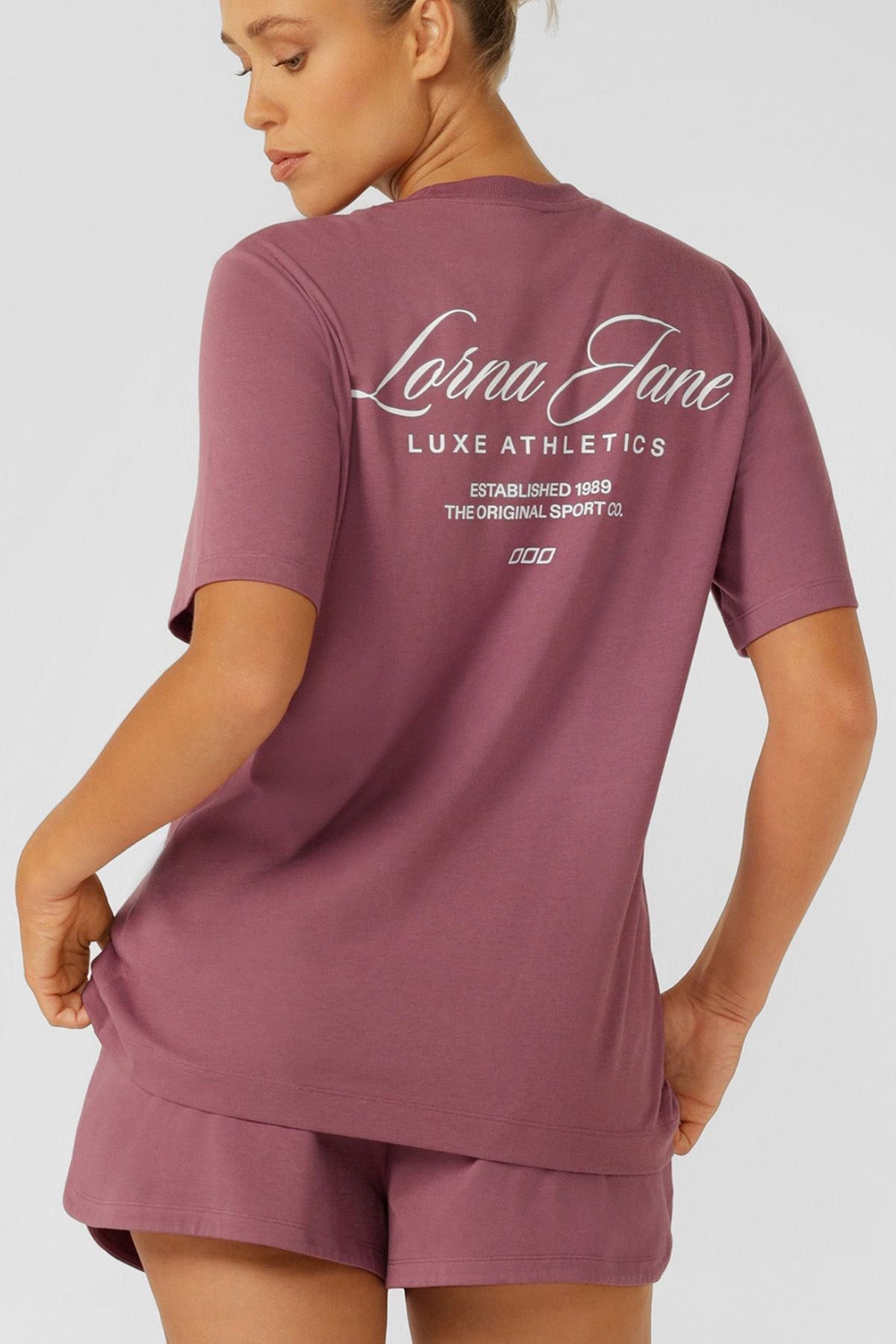 A woman faces away, wearing a mauve Lorna Jane Luxe Athletic Relaxed T-Shirt and matching shorts. The back of her shirt displays "Lorna Jane Luxe Athletics Established 1989 The Original Sport Co." in white text.