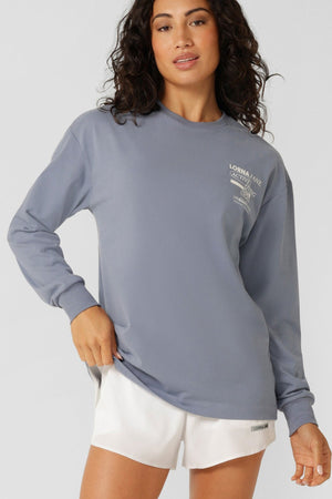 A woman with wavy dark hair wears a loose gray cotton Lorna Jane Momentum Plush Long Sleeve T-Shirt with white chest text and white athletic shorts, standing against a plain light background.