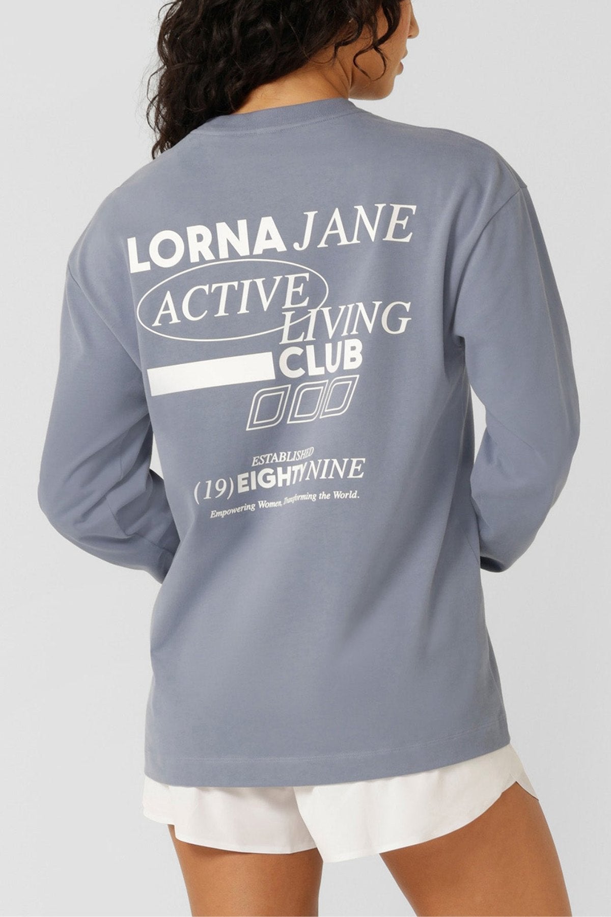 A woman with long, wavy hair faces away from the camera, wearing a light blue Lorna Jane Momentum Plush Long Sleeve T-Shirt with "Lorna Jane Active Living Club" graphics on the back and paired with white shorts.