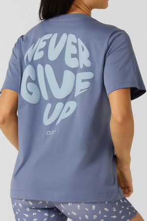 A person, turned away from the camera, wears a blue Lorna Jane Never Give Up Relaxed T-Shirt with “NEVER GIVE UP” in large light blue letters on the back and blue patterned shorts—ideal for an active lifestyle.