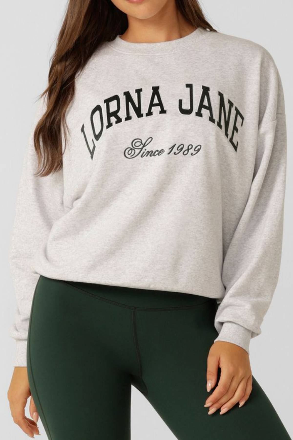 A person wears the Lorna Jane New Sport Sweatshirt in light gray cotton terry with “LORNA JANE Since 1989” in black, paired with dark green leggings, shown from neck to mid-thigh.