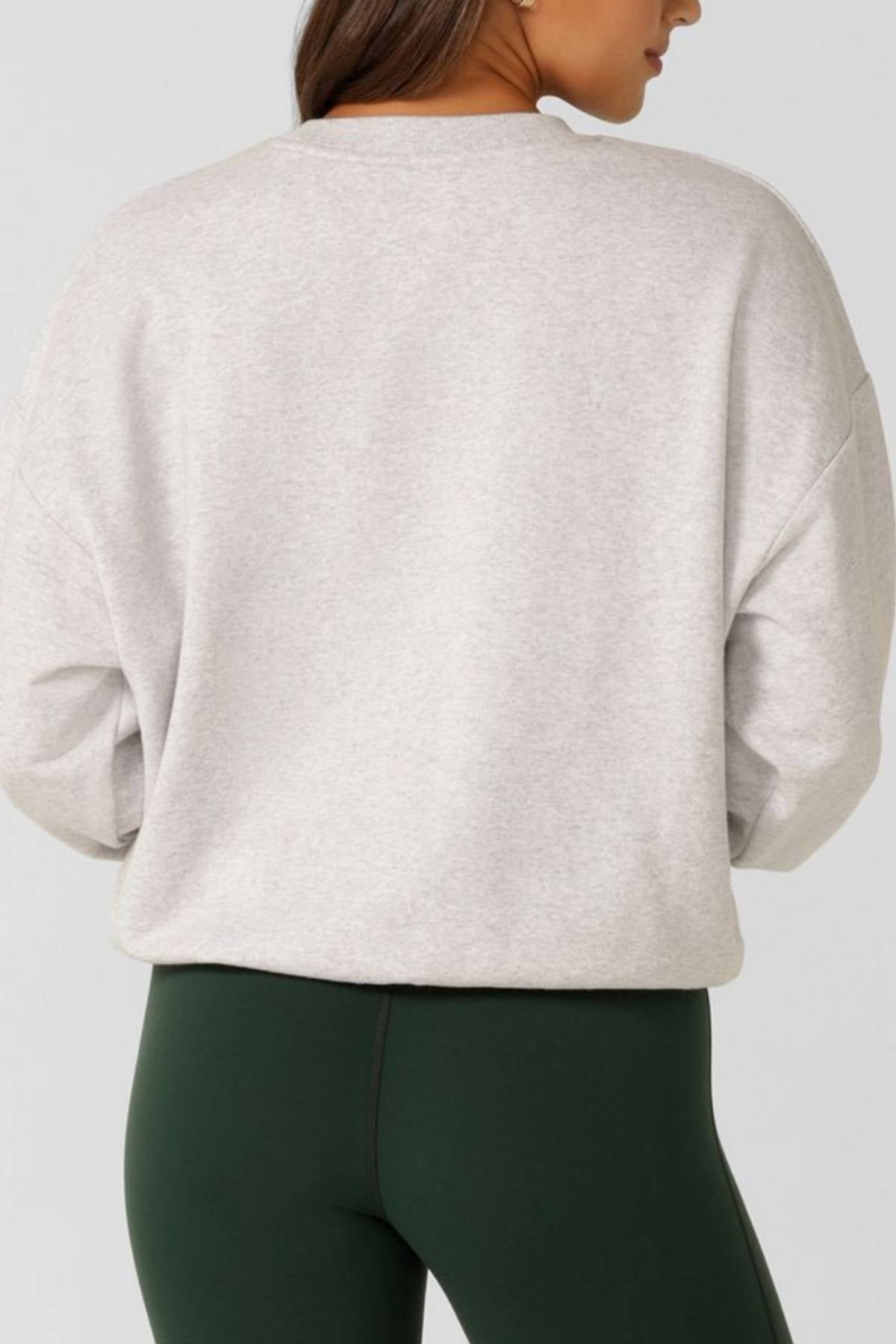A person with long brown hair is shown from the back, wearing the Lorna Jane New Sport Sweatshirt in an oversized fit and high-waisted dark green leggings against a plain background.