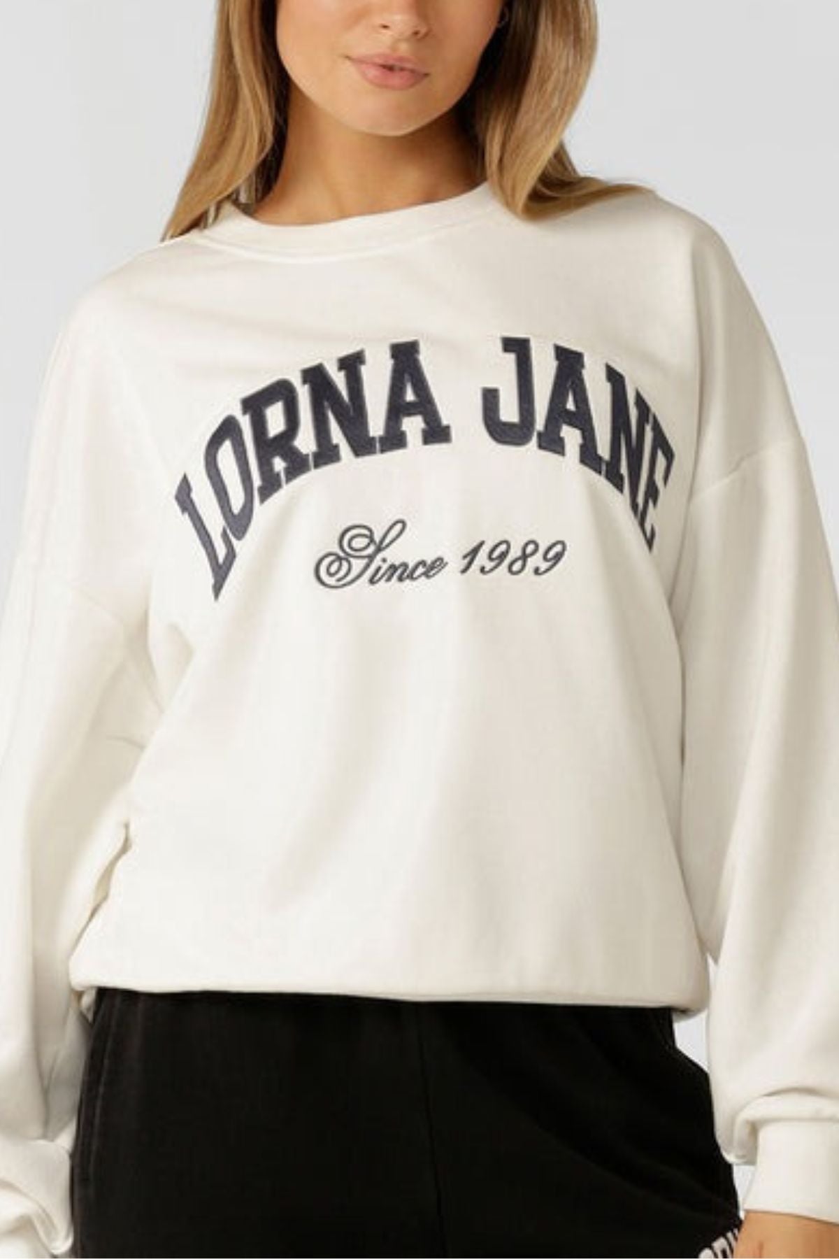 A woman wears the Lorna Jane New Sport Sweatshirt, an oversized white cotton terry top with "LORNA JANE Since 1989" in black, paired with black pants. Her face is partially visible.