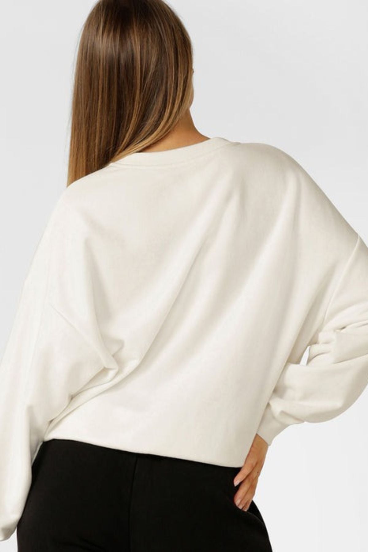 Seen from behind against a plain light background, a person with long straight hair wears the Lorna Jane New Sport Sweatshirt in cotton terry and black pants, with one hand resting on their hip.