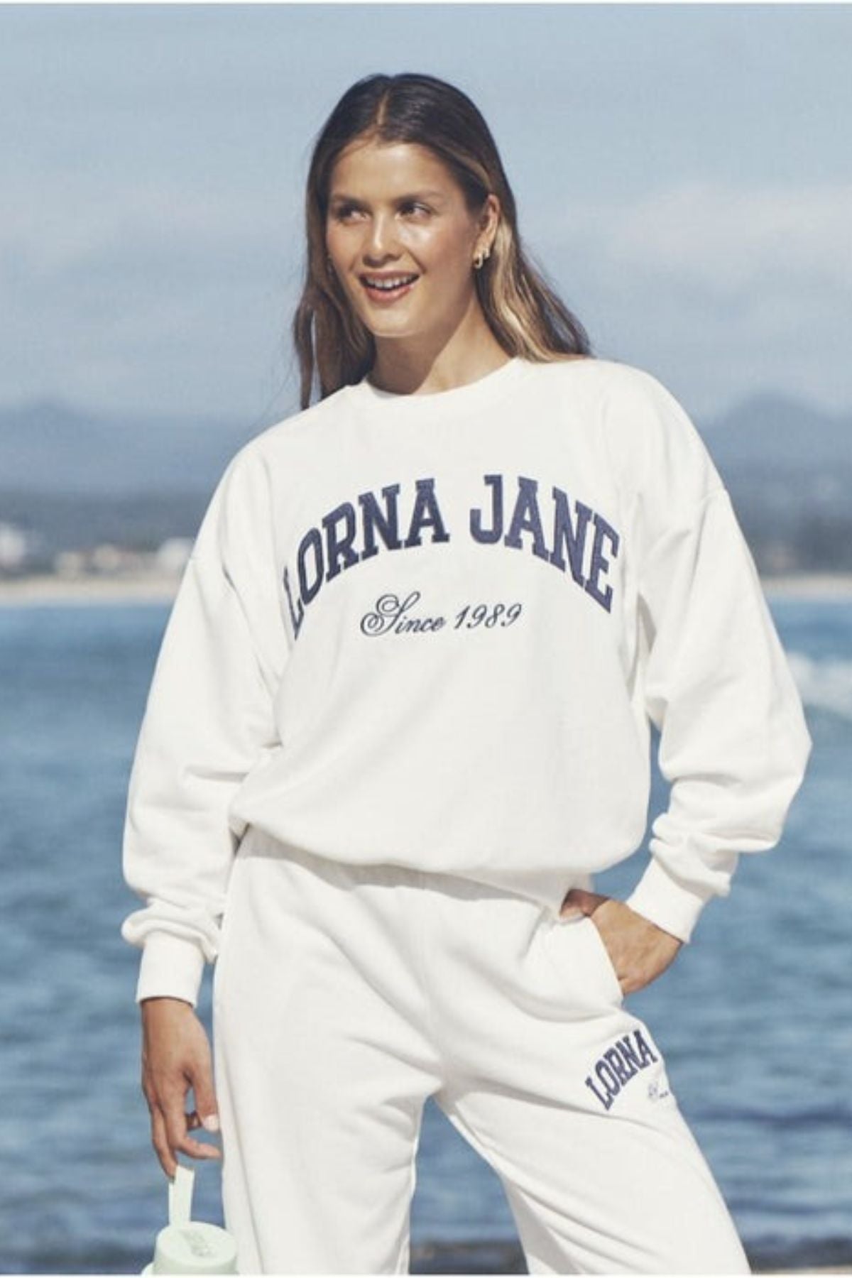 A woman smiles by the ocean wearing a Lorna Jane New Sport Sweatshirt and sweatpants with "Lorna Jane Since 1989" on the top, while holding a light green water bottle.