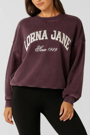 A woman wears a purple Lorna Jane New Sport Washed Sweatshirt with "LORNA JANE Since 1989" in white, paired with black leggings. Her face is partially out of frame.