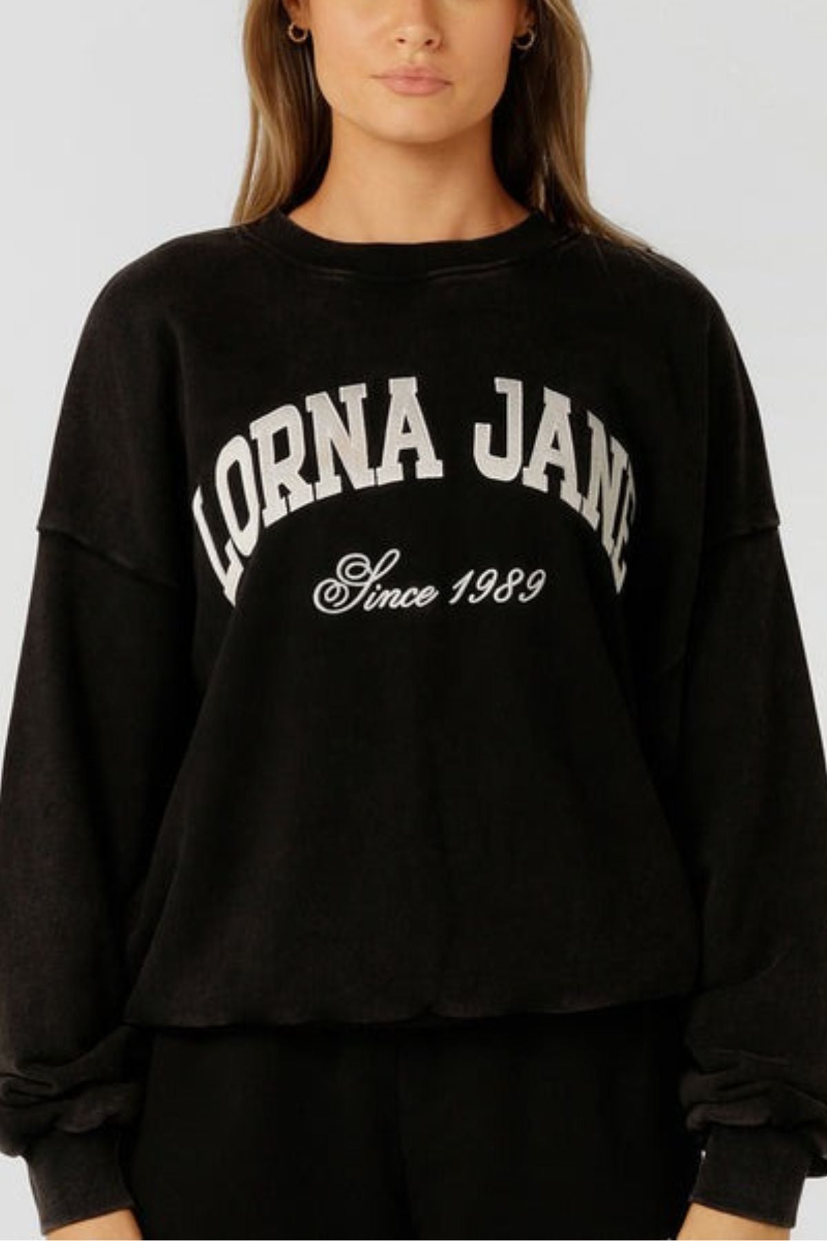 A woman wears the Lorna Jane New Sport Washed Sweatshirt—an oversized, vintage-inspired black cotton terry pullover with "LORNA JANE" and "Since 1989" printed in white. A simple backdrop highlights this classic sporty design.