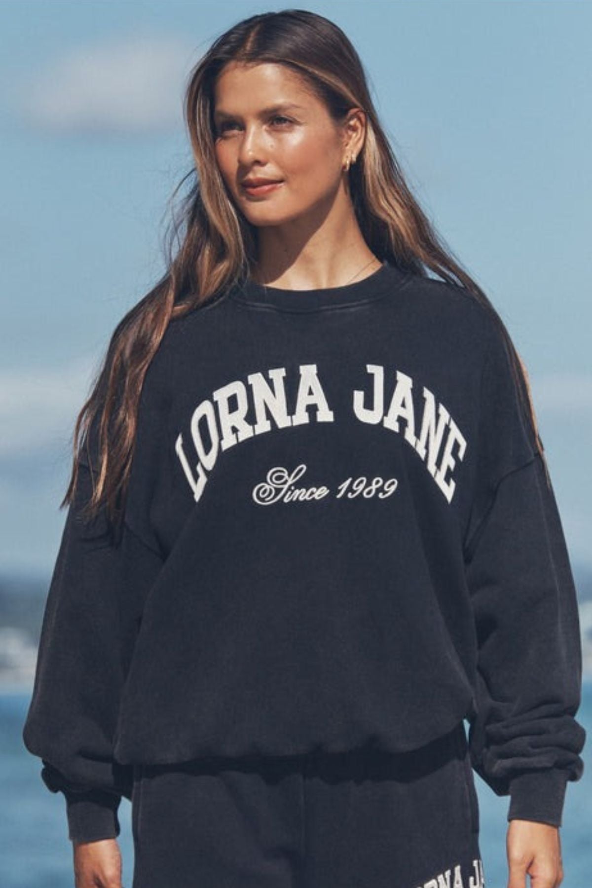 A woman with long brown hair stands outdoors in a black Lorna Jane New Sport Washed Sweatshirt with an oversized fit, set against a blurred background featuring hints of blue sky and clouds.