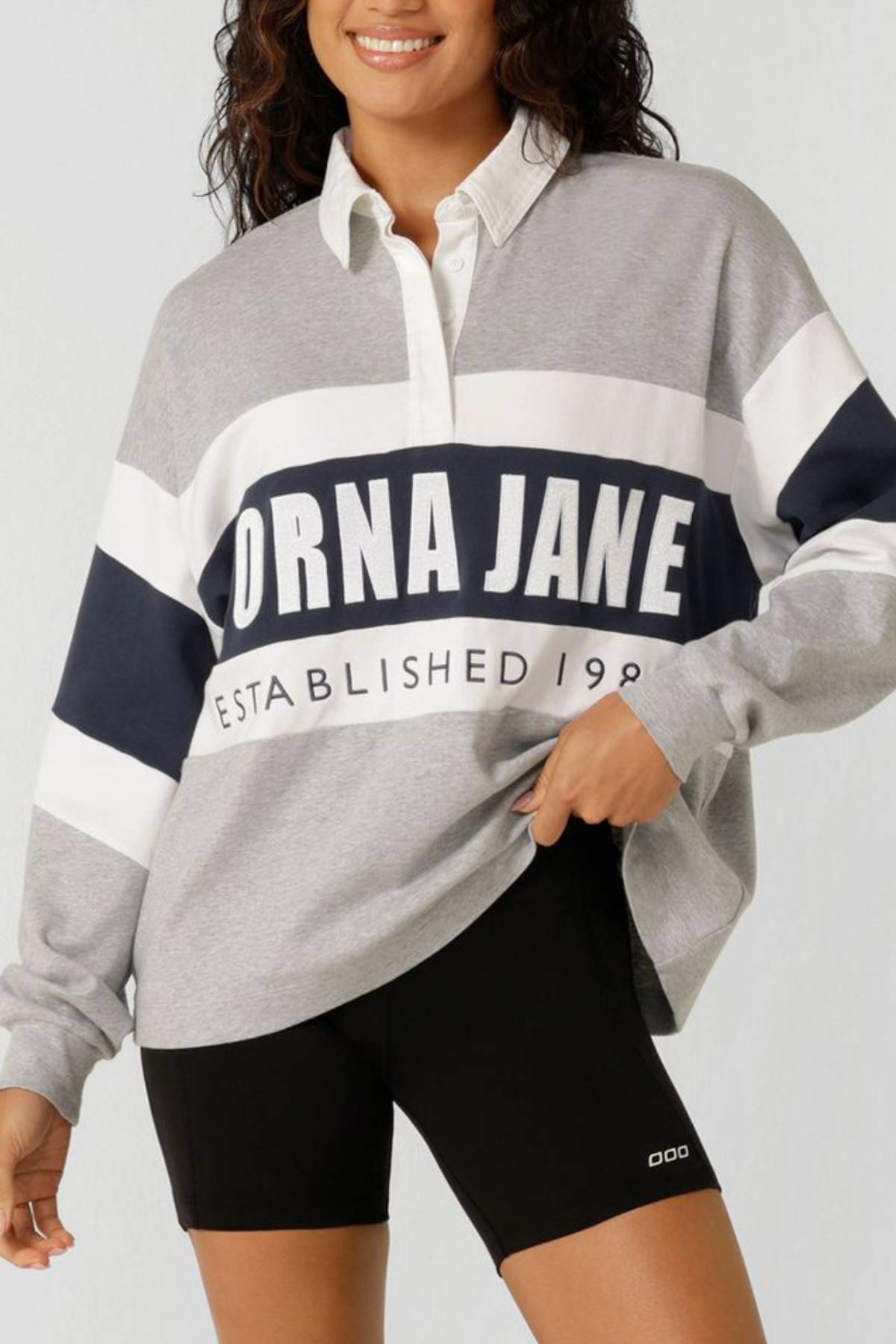 A woman models the Lorna Jane Original Sport Rugby Sweat in grey, white, and navy with "LORNA JANE ESTABLISHED 1989" text, paired with black athletic shorts for a sporty athleisure look.