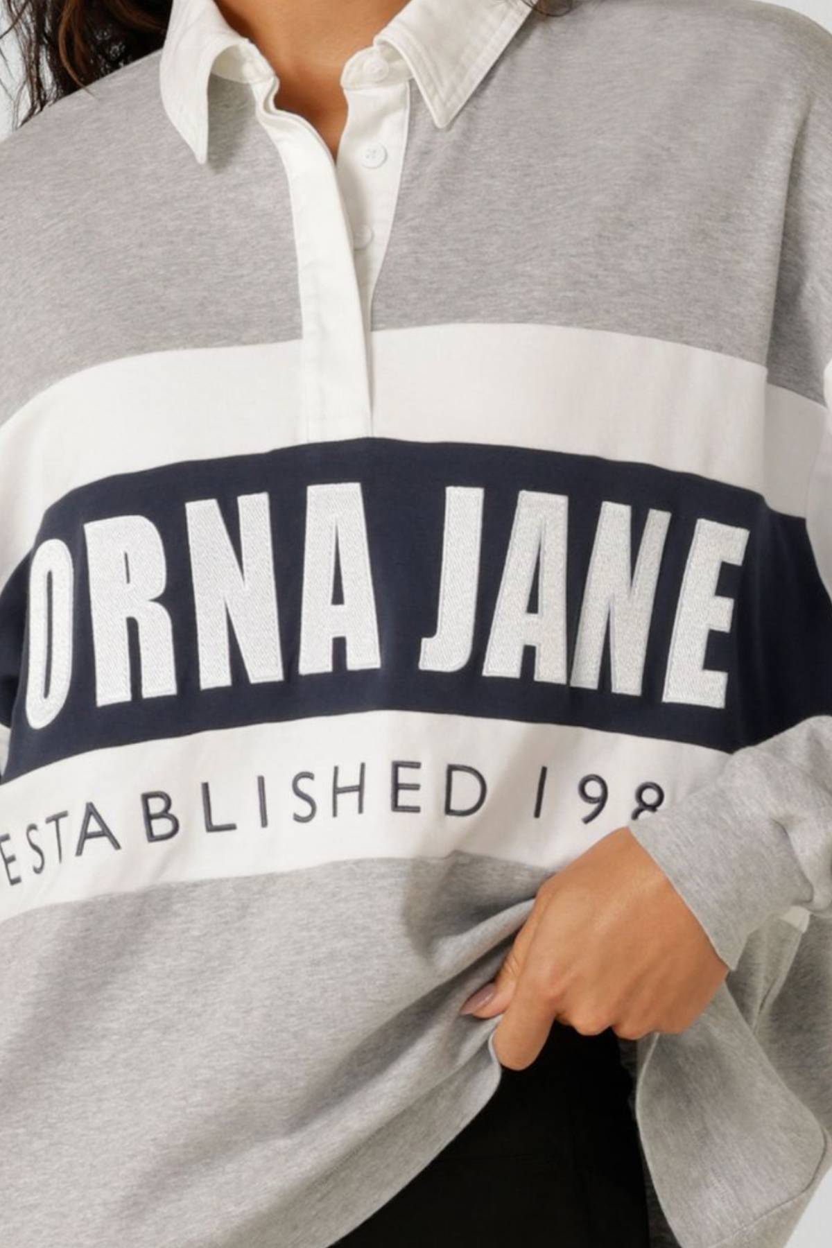 Wearing the Lorna Jane Original Sport Rugby Sweat in gray, white, and navy with "LORNA JANE ESTABLISHED 1989" on the front, a person displays classic athleisure style while holding the hem of their sweatshirt.