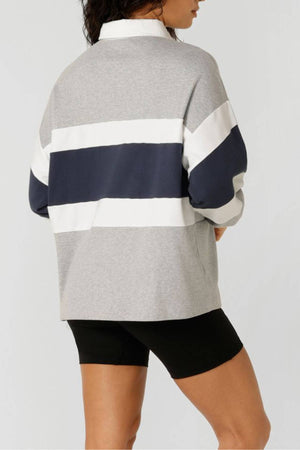 A person is seen from behind wearing the Lorna Jane Original Sport Rugby Sweat in gray with white and navy stripes, paired with black cycling shorts. The plain light background highlights their athleisure style.