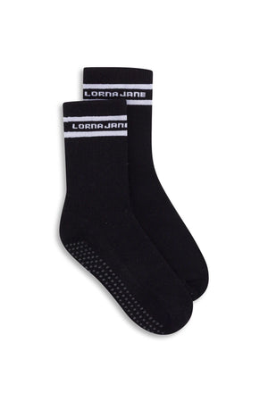 The Lorna Jane Pilates Grip Crew Socks are black crew socks featuring a white stripe and "LORNA JANE" text at the top. Designed specifically for Pilates, these socks have a grippy sole with small non-slip dots on the bottom to provide enhanced stability and support.