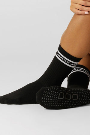 A person wearing Lorna Jane Pilates Grip Crew Socks—black with white trim and text—sits with one knee up, hands resting on their leg. The crew socks feature rubber grips on the sole for added non-slip stability.