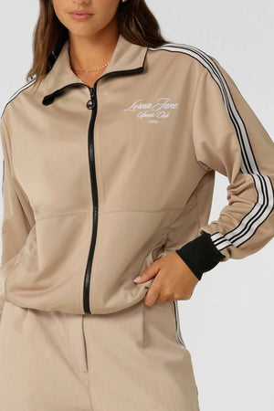 A woman wears the Lorna Jane Retro Track Zip Through Jacket in beige with black and white striped sleeves, zip pockets, matching pants, and "Lorna Jane Sports Club 1990" in white on the chest.