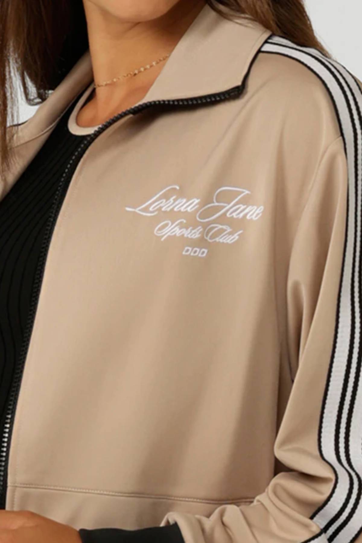 A woman wears the Lorna Jane Retro Track Zip Through Jacket in beige, featuring black and white striped sleeves, "Lorna Jane Sports Club" embroidery on the chest, and an athletic fit for comfort.