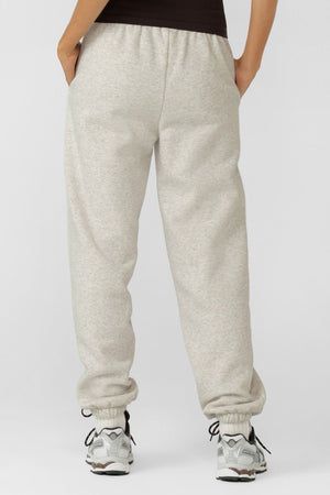 Someone in relaxed fit Lorna Jane Sport Mode Sweatpants made from cotton blend fleece, white sneakers, and a dark top stands facing away with hands in pockets against a plain light background.