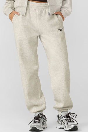 Someone wears relaxed fit, light beige Lorna Jane Sport Mode Sweatpants with hands in pockets, styled with a matching cotton-blend fleece sweatshirt and white athletic sneakers with black laces, standing against a plain light backdrop.