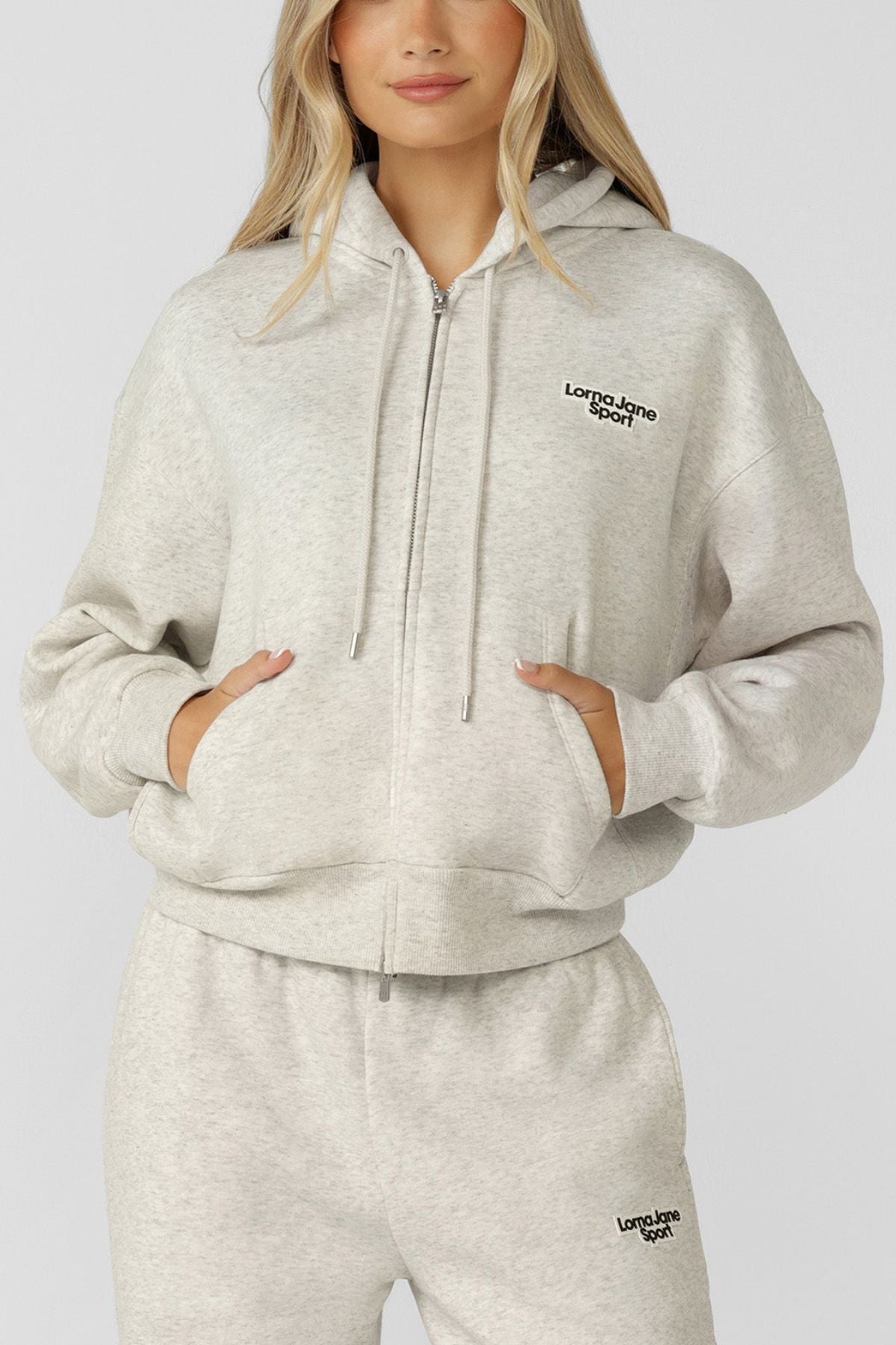A woman stands against a plain background wearing the Lorna Jane Sport Mode Zip Through Hoodie in light grey cotton-blend fleece, featuring "Lorna Jane Sport" logos and an adjustable drawcord hood, with her hands in her pockets.