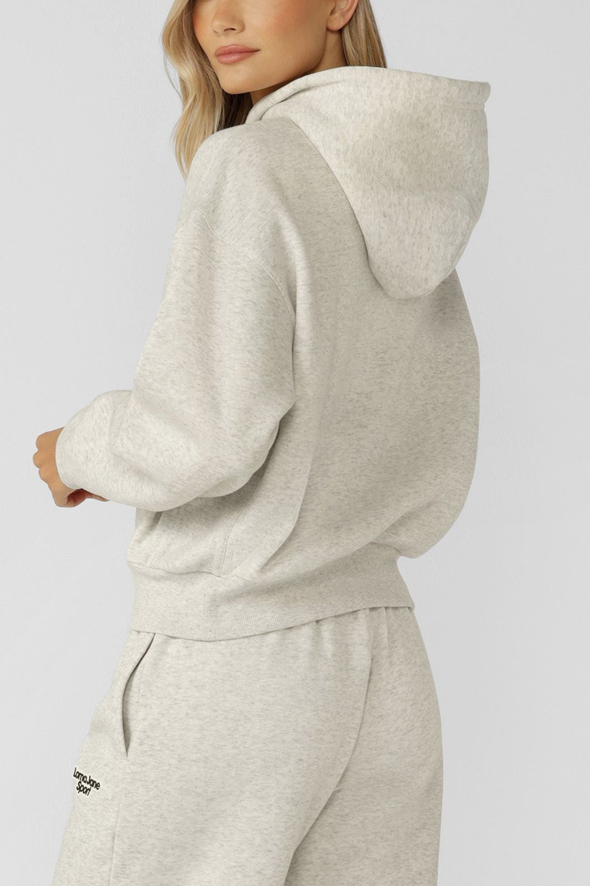 A woman with long blonde hair wears the Lorna Jane Sport Mode Zip Through Hoodie in light gray cotton blend fleece and matching sweatpants, standing sideways and looking over her shoulder against a plain light background.