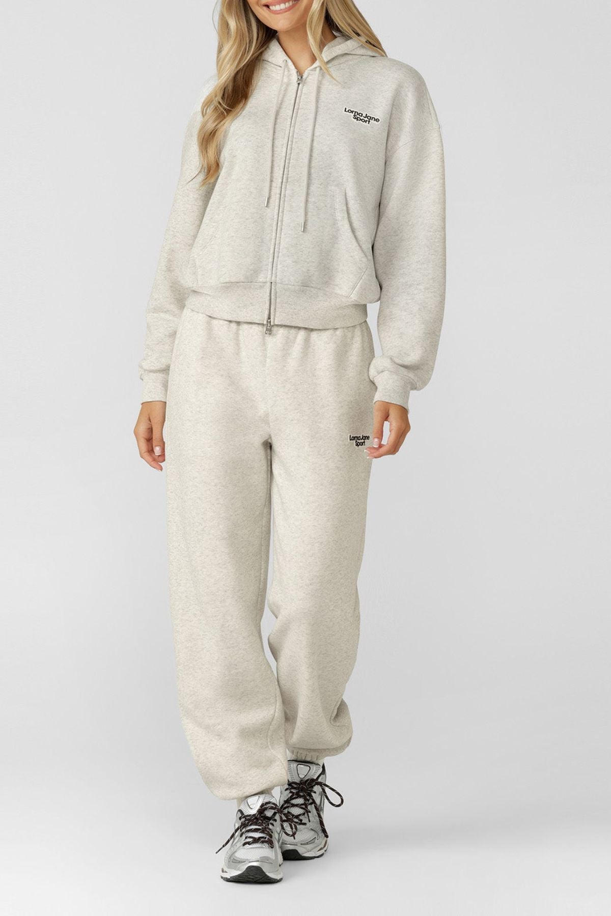 A person wears the Lorna Jane Sport Mode Zip Through Hoodie in light grey with matching sweatpants and white-grey sneakers. The tracksuit shows small black text on the chest and hip, set against a plain background.