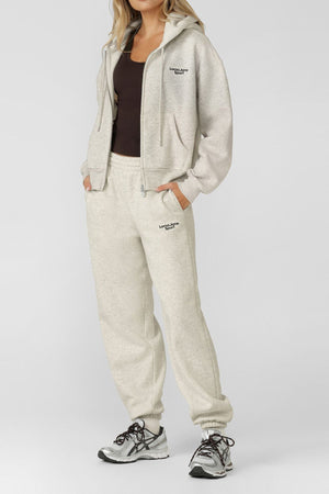 A woman wearing a Lorna Jane Sport Mode Zip Through Hoodie in light gray with black text, matching sweatpants, a dark brown tank top, and gray sneakers stands against a plain light background.