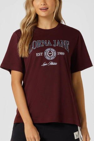 A woman with long, wavy blonde hair wears a maroon Lorna Jane Strive Relaxed T-Shirt featuring "LORNA JANE EST 1989" and "Love Athlete" on the front.