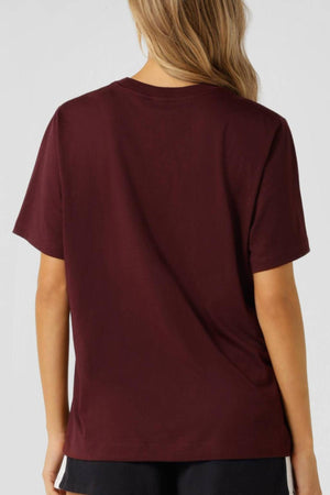 A person with long, wavy blonde hair is shown from the back, wearing the Lorna Jane Strive Relaxed T-Shirt and black pants against a light gray background.