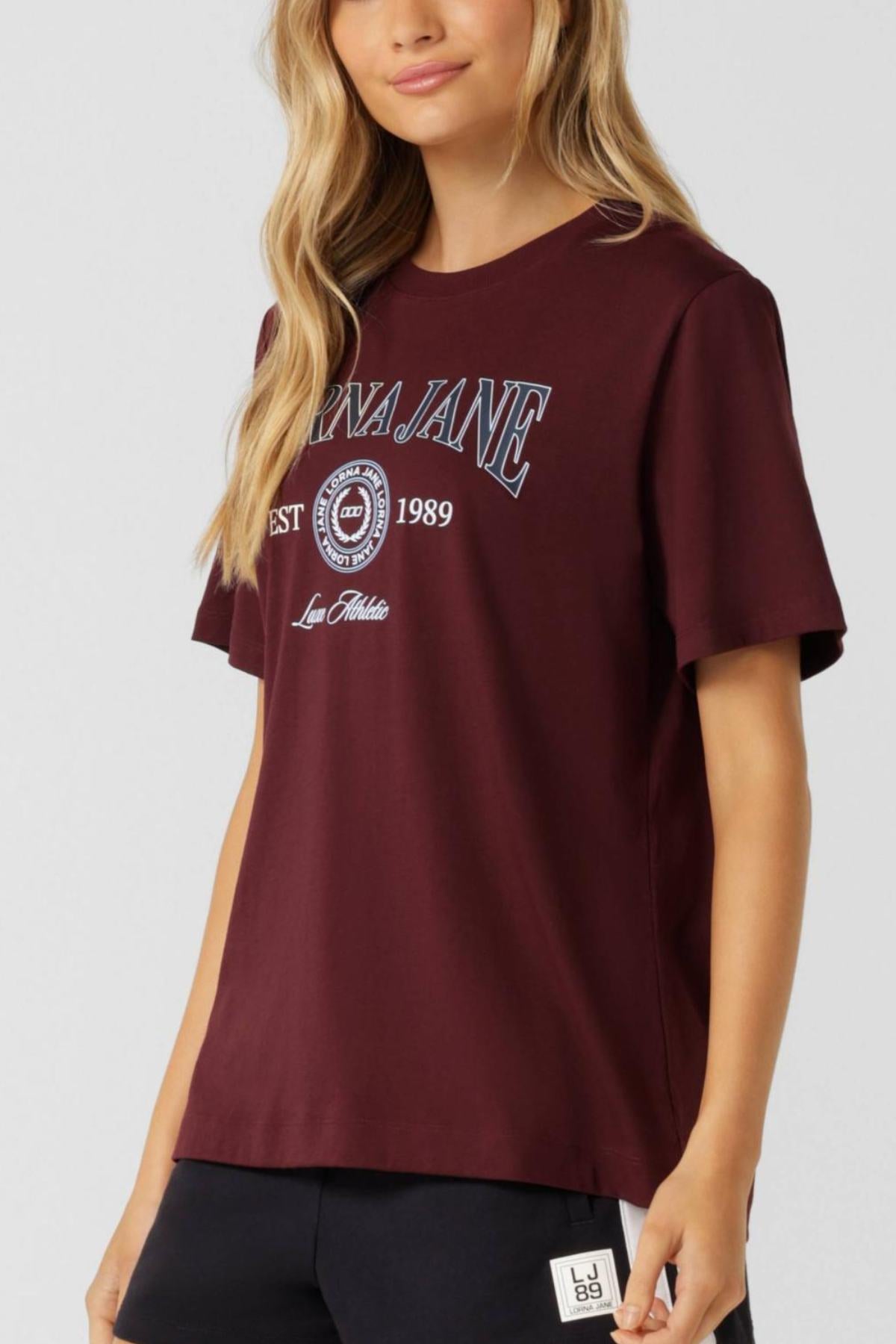 A woman wears the Lorna Jane Strive Relaxed T-Shirt in burgundy cotton blend with "Lorna Jane Est 1989" and a circular logo on the front, styled with black shorts against a plain light background.