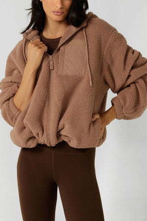 A woman stands with one hand on her hip, wearing the Lorna Jane Teddy Zip Thru Hoodie in brown—an ultra-soft hoodie with a chest pocket—paired with matching brown leggings.