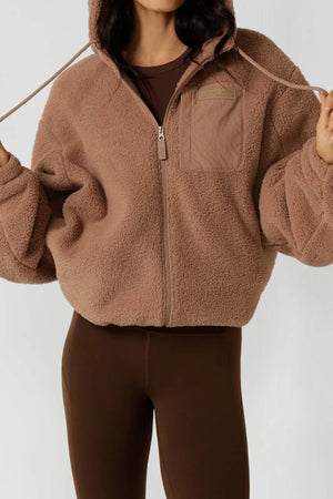 Wearing the Lorna Jane Teddy Zip Thru Hoodie in brown, featuring an adjustable hood, chest pocket, and ultra-soft fabric, with drawstrings in hand and paired with matching leggings. The person’s face is not shown.
