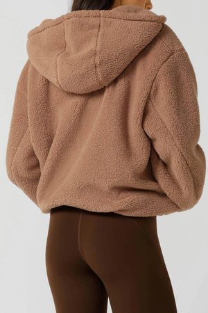 A person is seen from the back wearing a brown Lorna Jane Teddy Zip Thru Hoodie with an adjustable hood and dark brown leggings against a plain background.