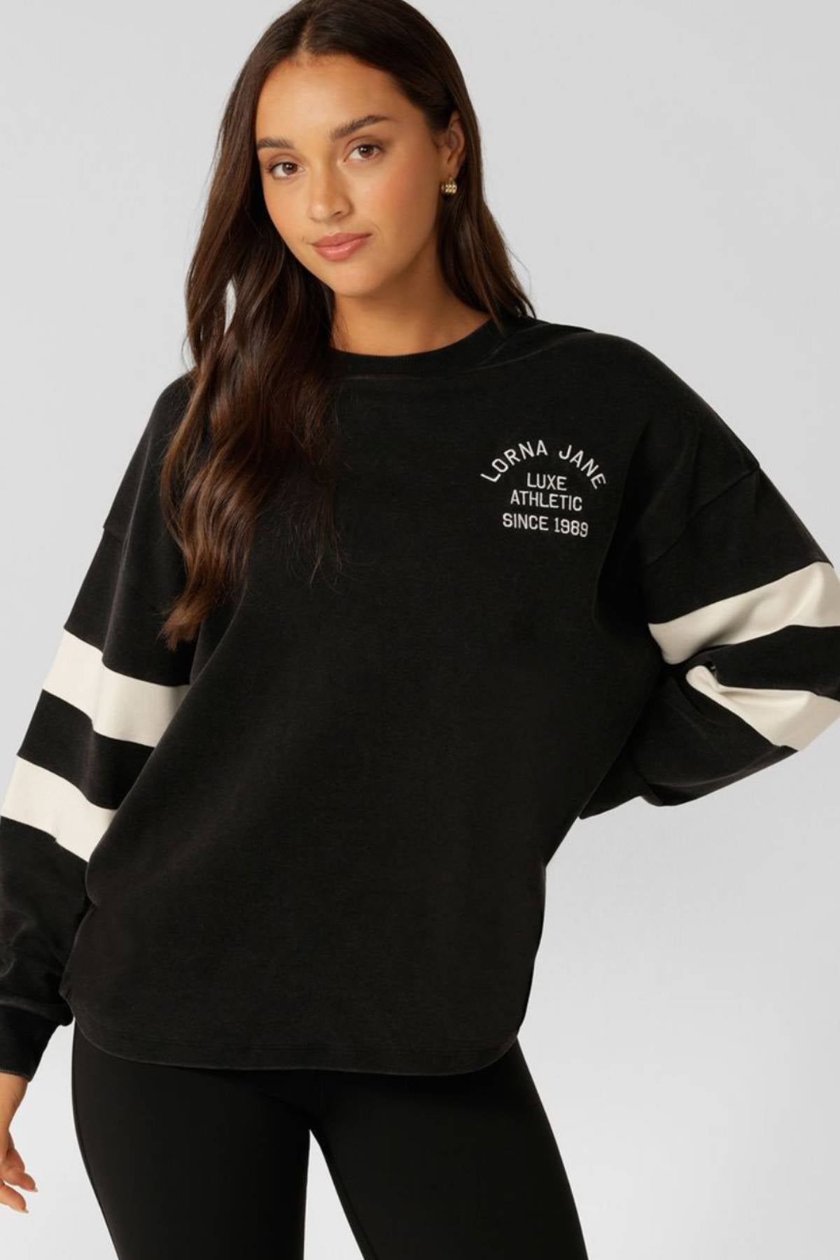 A woman with long brown hair wears the Lorna Jane Throwback Oversized Lightweight Sweat with white sleeve stripes and chest text, paired with black leggings, standing against a light gray background.